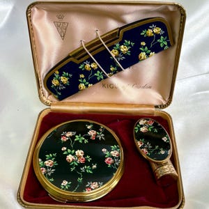 May include: A vintage vanity set presented in a gold-toned case. The set features a comb, compact, and hand mirror, each with a dark blue floral design of yellow and red roses. The case is lined with pink satin and the text "KIGU of London".