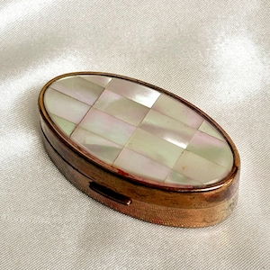 Könnte beinhalten: Eine ovale, antike Pillendose mit einem karierten Deckel aus Perlmutt und einem bronzefarbenen Metallrahmen. Die geschlossene Dose liegt auf einer weißen, strukturierten Oberfläche. Das Perlmutt hat einen perlmuttartigen Glanz.
