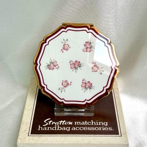 May include: A vintage Stratton compact mirror with a white surface, adorned with pink floral designs. The compact has a burgundy border and gold trim. The box below the compact reads "Stratton matching handbag accessories."