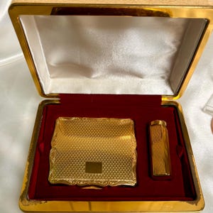May include: A vintage gold-toned compact and lighter set in a rectangular box. The compact and lighter have a textured design. The box is gold-toned with a red velvet interior and a cream-colored satin lining.