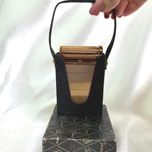 Op de afbeelding: Een vintage goudkleurige compacte spiegel met een zwarte draagtas en handvat. De compacte spiegel heeft een rechthoekige vorm met een gaasfront. De etui staat op een doos met patroon.