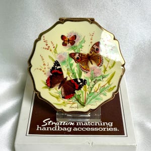 May include: A round, scalloped-edge compact mirror with a cream-colored background featuring three butterflies and thistle flowers. The gold-tone metal frame surrounds the artwork. It rests on a white box labeled Stratton matching handbag accessories.