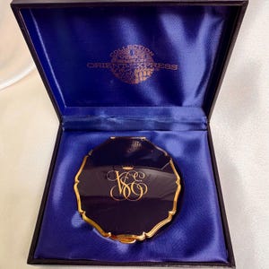 May include: A navy blue compact mirror with gold trim and a gold monogram, presented in a blue velvet-lined box. The box is embossed with "COLLECTION VENICE SIMPLON ORIENT EXPRESS" in gold lettering.
