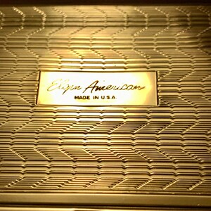 May include: A gold-colored compact with a geometric pattern and the text "Elgin American Made in U.S.A."