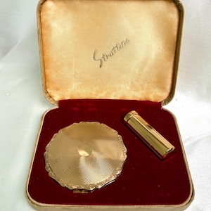 May include: A vintage gold-coloured Stratton compact and lipstick holder set. The compact features a detailed, patterned design and a scalloped edge. The set is presented in a beige and red velvet lined case, with the name "Stratton" printed inside.