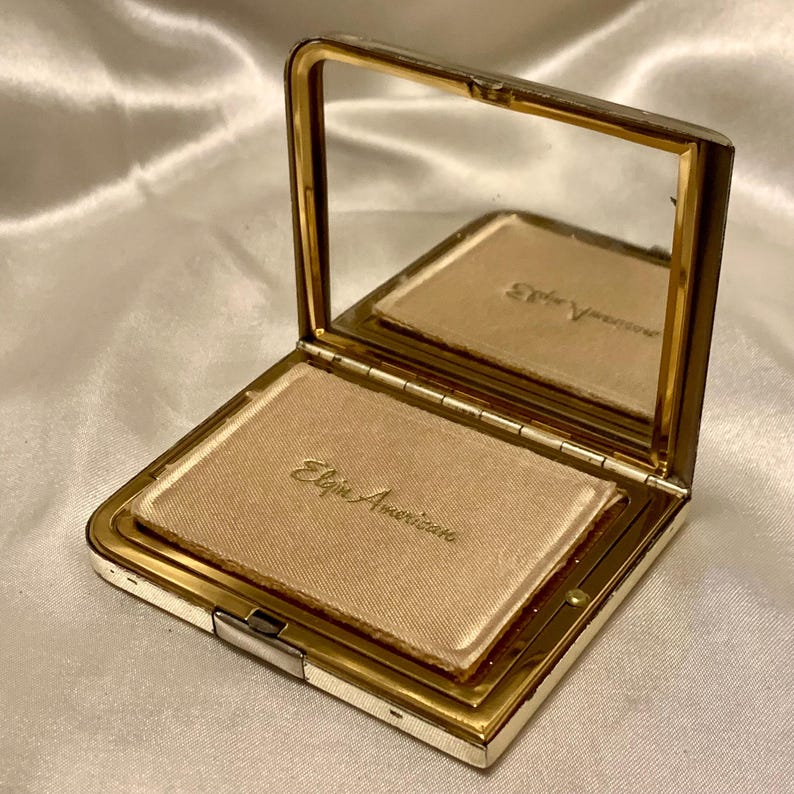 May include: A gold-colored compact with a hinged mirror and a white satin interior. The interior is labeled "Elgin American".