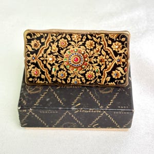 May include: A vintage rectangular compact featuring a black and gold floral pattern, embellished with colourful gemstones. It rests on a black box with the text "Stratton" and "Made in England". The compact's detailed design and bright colours make it a decorative item.