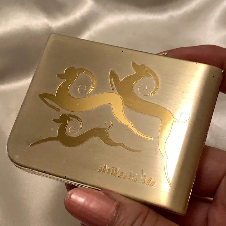May include: A silver compact with a gold design of three stylized deer. The compact has the words "New York" engraved on the bottom.