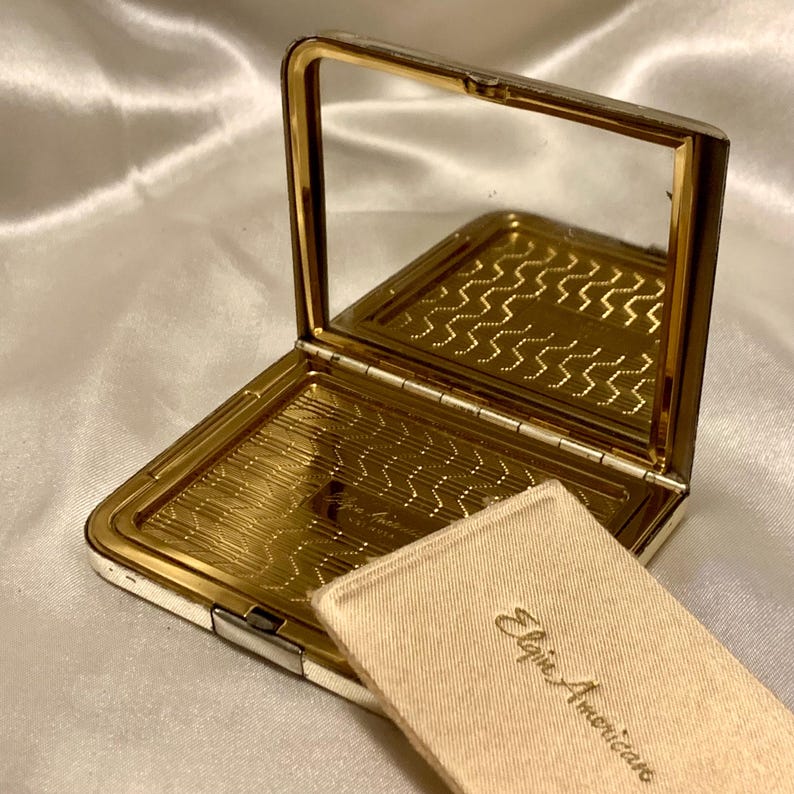May include: A silver and gold compact mirror with a white satin pouch. The pouch has the text "Elsie American" printed on it.