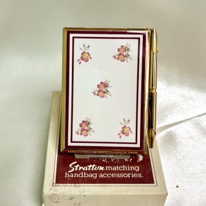 May include: A gold-toned compact mirror with a white face featuring a floral design in pink and yellow. The compact has a burgundy border and is on a box with the text "Stratton matching handbag accessories."
