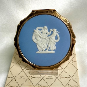 May include: A round gold-tone metal compact mirror featuring a scalloped edge and a light blue circular inlay. The center displays a white relief of three figures dancing in flowing garments. The brand name Stratton is visible on the base.