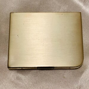 May include: A silver metal compact with a smooth, brushed finish. The compact is rectangular with rounded corners.