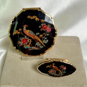 May include: A round gold-rimmed compact mirror and a smaller oval gold-rimmed compact mirror, both featuring a black enamel surface with a colorful illustration of a pheasant perched on a flowering branch with red blossoms and green leaves.
