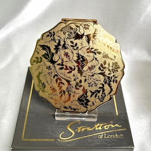 May include: A round gold-tone metal compact mirror with a scalloped edge and an intricate black floral and leaf pattern engraved on the lid. The compact rests on a dark gray box featuring the gold-printed text Stratton of London on the front.