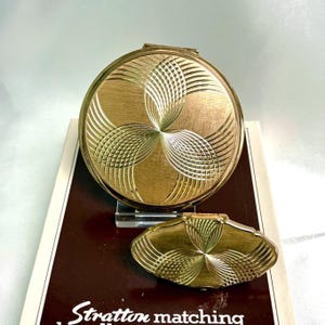 May include: A vintage gold-toned compact and a matching small case. Both have a textured, floral-like design. The compact is round, and the small case is oval-shaped. The text "Stratton matching" is visible on the packaging.