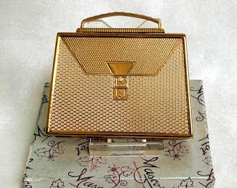 May include: A gold-toned metal compact case shaped like a miniature briefcase with a textured surface, a top handle, and a central latch detail. The compact rests on a white box featuring the brand name Mascot London in a decorative script font.