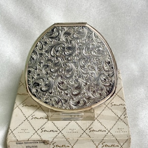 May include: A silver-toned metal compact mirror with a rounded triangular shape and an intricate embossed scrollwork pattern on the lid. The compact rests on a white cardboard box featuring a repeating diamond pattern and the text Stratton Made in England.