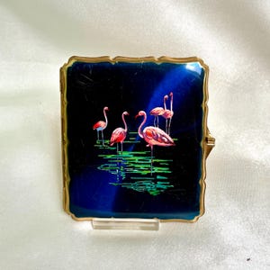 May include: A square metal compact case with a gold-toned scalloped frame. The front features a deep blue enamel surface decorated with a painted scene of four pink flamingos standing in green water, with a subtle light reflection across the center.