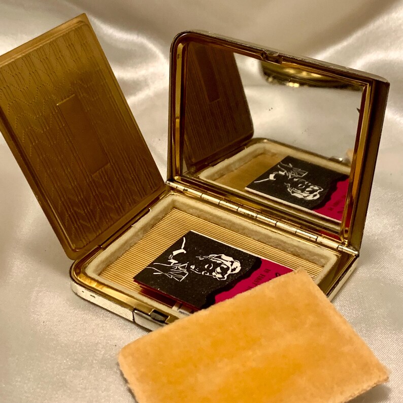 May include: A gold compact mirror with a hinged lid and a mirror inside. The compact has a white velvet lining and a small, black and white image of a woman on the inside. The compact is open and a small, orange velvet cushion is sitting next to it.