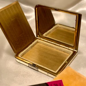 May include: A vintage gold compact with a mirror and a white interior. The compact is open and the mirror is visible. The compact is in good condition and has a few scratches.