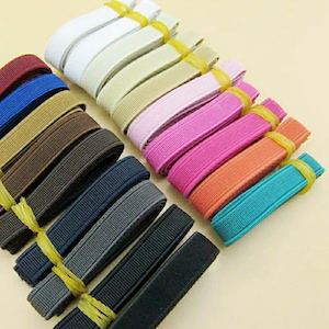 Pode incluir: Conjunto de 12 cores de fita elástica, cada cor é um tom diferente de marrom, azul, cinza, rosa, laranja e branco. A fita está disposta em forma de leque, com as cores mais escuras na parte inferior e as cores mais claras na parte superior.