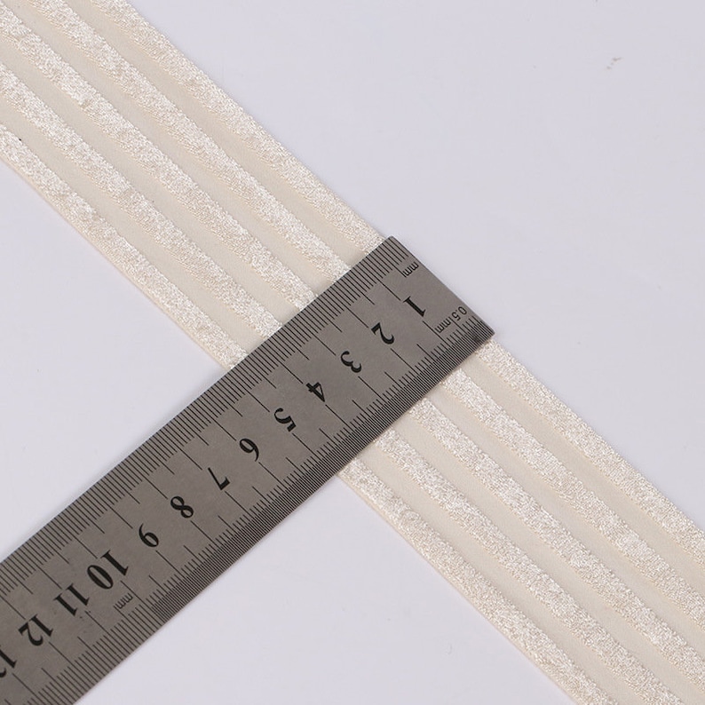Peut inclure: Un ruban en tissu de couleur cr&egrave;me orn&eacute; de trois bandes parall&egrave;les textur&eacute;es et scintillantes dispos&eacute;es dans le sens de la longueur. Une r&egrave;gle en acier inoxydable est pos&eacute;e sur le ruban, indiquant une largeur d'environ 3,8 cm.