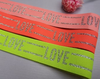 Könnte beinhalten: Drei Gummibänder mit dem Wort "LOVE" in silbernen Glitzer auf jedem Band. Die Bänder sind rosa, orange und grün.