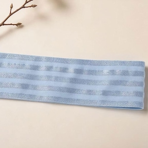 Peut inclure: Un bandeau &eacute;lastique bleu clair pr&eacute;sentant un motif r&eacute;p&eacute;titif de rayures horizontales. Le tissu poss&egrave;de une texture lisse avec une alternance de rayures mates et de rayures aux reflets m&eacute;talliques sur toute la longueur de la bande.