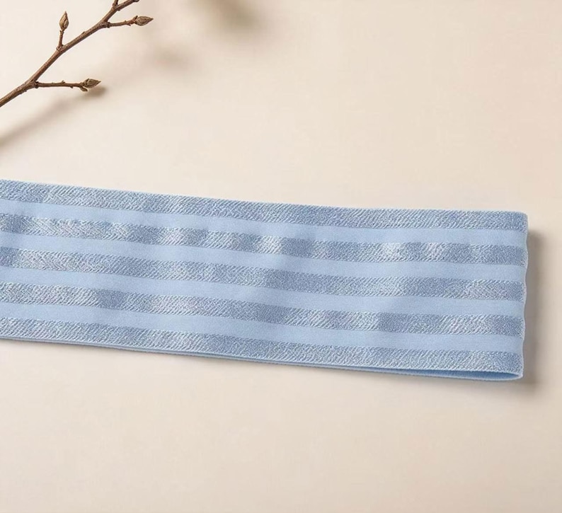 Peut inclure: Un bandeau &eacute;lastique bleu clair pr&eacute;sentant un motif r&eacute;p&eacute;titif de rayures horizontales. Le tissu poss&egrave;de une texture lisse avec une alternance de rayures mates et de rayures aux reflets m&eacute;talliques sur toute la longueur de la bande.