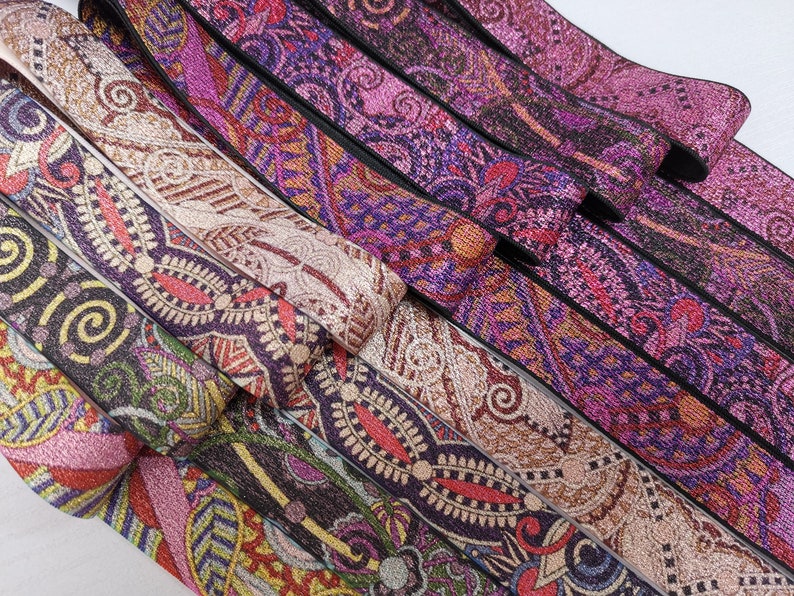 May include: A collection of colourful patterned elastic bands with a glittery finish. The bands are arranged in a stack, with the top band being a light brown with a gold glitter finish. The other bands are a mix of purple, pink, and black with various patterns.