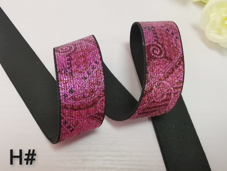 May include: Pink elastic trim with a black backing. The trim has a metallic sheen and a floral pattern.