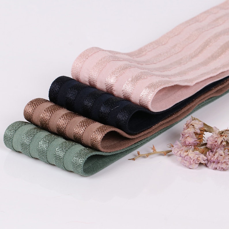 Peut inclure: Une pile de quatre bandes &eacute;lastiques pli&eacute;es en rose clair, noir, bronze et vert sauge. Chaque bande pr&eacute;sente un motif r&eacute;p&eacute;titif de rayures horizontales mates et m&eacute;talliques. Les bandes sont superpos&eacute;es, montrant leur texture tiss&eacute;e.
