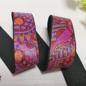 May include: A close-up of a black elastic band with a pink, orange and purple patterned trim. The trim has a metallic sheen. The elastic band is folded in half, showing both sides of the trim.
