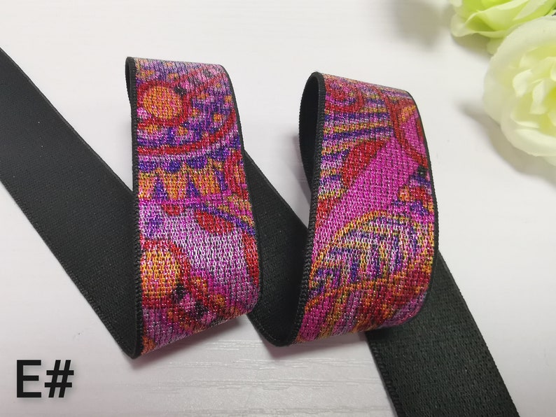 May include: A close-up of a black elastic band with a pink, orange and purple patterned trim. The trim has a metallic sheen. The elastic band is folded in half, showing both sides of the trim.
