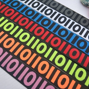May include: Seven different colors of elastic trim with a repeating pattern of black circles and the text "10110110110110". The colors are pink, orange, lime green, red, blue, white, and gray.