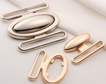 May include: An assortment of metal hardware components in silver and gold tones. The collection includes oval-shaped buckles, rectangular loops, and decorative elements, all with a polished finish. The pieces are arranged on a white fabric background.