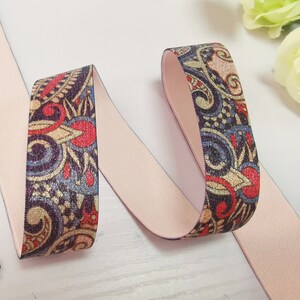 May include: A close-up of a pink elastic band with a paisley pattern in shades of blue, gold, and red. The elastic band is folded in half, and the patterned side is facing the camera.