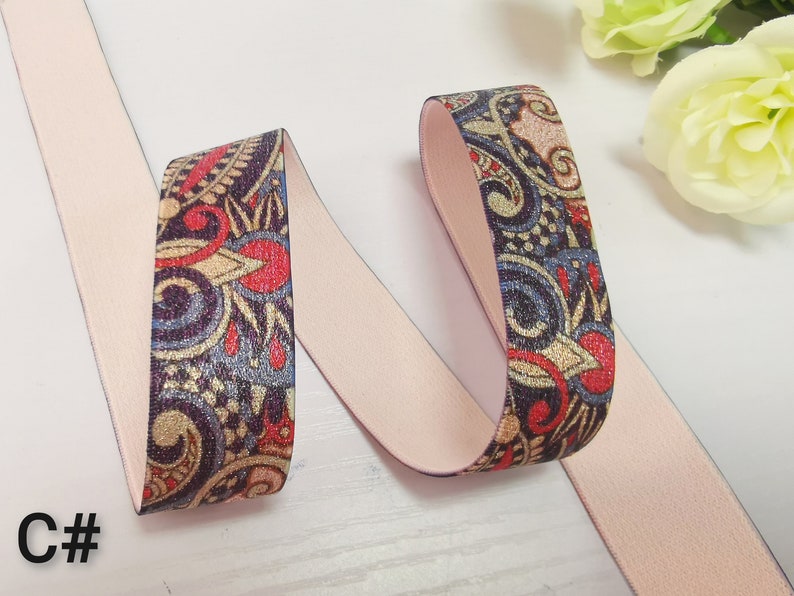 May include: A close-up of a pink elastic band with a paisley pattern in shades of blue, gold, and red. The elastic band is folded in half, and the patterned side is facing the camera.