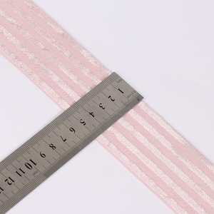 Peut inclure: Un ruban rose clair orn&eacute; d'une s&eacute;rie de rayures verticales scintillantes. Une r&egrave;gle en acier inoxydable est plac&eacute;e sur le ruban, indiquant une largeur d'environ 3,8 cm. Le ruban pr&eacute;sente une texture lisse et semi-transparente avec des reflets m&eacute;talliques.