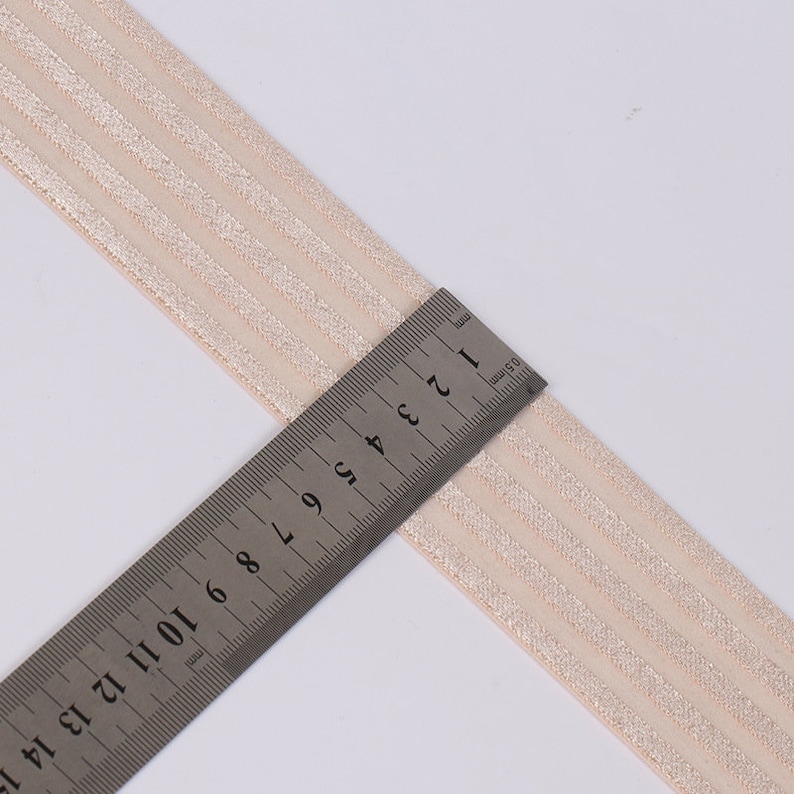 Peut inclure: Une bande &eacute;lastique beige clair avec quatre rayures scintillantes parall&egrave;les. Une r&egrave;gle m&eacute;tallique est plac&eacute;e sur la largeur de la bande, indiquant une mesure d'environ 3,8 cm, ce qui correspond &agrave; la largeur totale de la bande de tissu.
