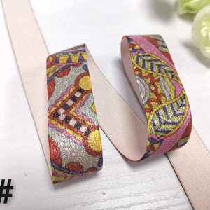May include: Two pieces of elastic trim with a pink and white background and a colourful paisley pattern. The trim is glittery and has a metallic sheen.