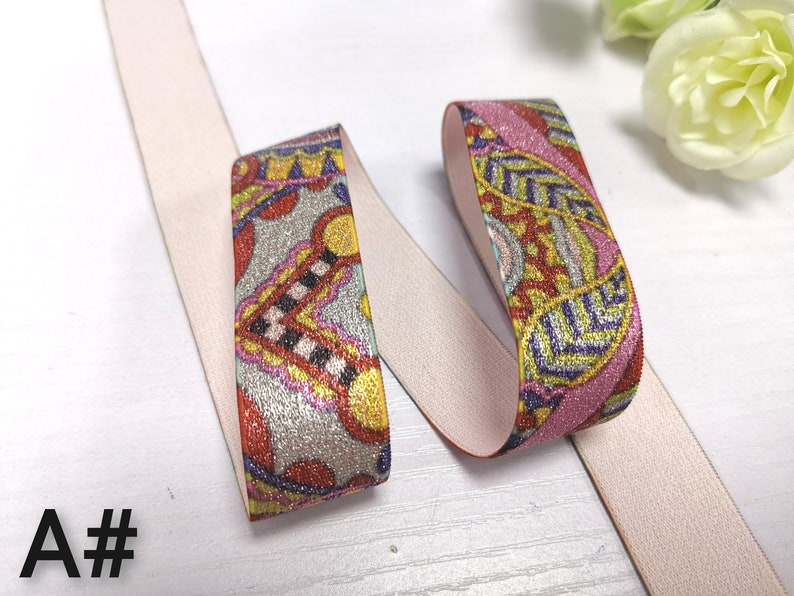 May include: Two pieces of elastic trim with a pink and white background and a colourful paisley pattern. The trim is glittery and has a metallic sheen.