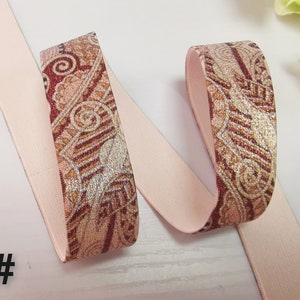 May include: Pink elastic ribbon with a brown and gold paisley pattern. The ribbon is folded in half and the patterned side is facing up.