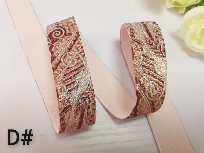 May include: Pink elastic ribbon with a brown and gold paisley pattern. The ribbon is folded in half and the patterned side is facing up.