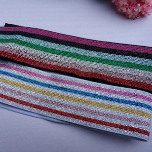 May include: Two rolls of elastic trim with a striped pattern. The trim is white with silver glitter and has stripes of pink, red, blue, and gold. The top roll has stripes of black, pink, silver, green, and red.