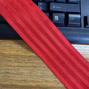 Peut inclure: Une bande &eacute;lastique rouge vif pr&eacute;sentant une s&eacute;rie de rayures textur&eacute;es en relief parall&egrave;les sur toute sa longueur. Le mat&eacute;riau a un aspect tiss&eacute; coh&eacute;rent avec un l&eacute;ger &eacute;clat, mettant en valeur une couleur uniforme et une conception structur&eacute;e.