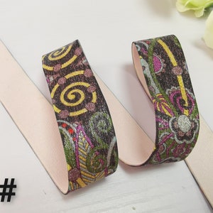 May include: Two pieces of elastic trim with a pink backing and a colourful patterned design. The pattern features swirls, flowers, and geometric shapes in shades of brown, yellow, green, pink, and silver.
