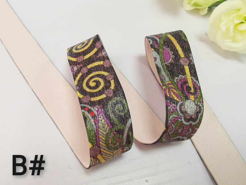 May include: Two pieces of elastic trim with a pink backing and a colourful patterned design. The pattern features swirls, flowers, and geometric shapes in shades of brown, yellow, green, pink, and silver.
