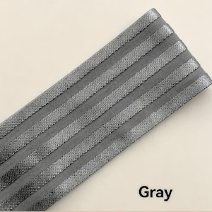 Peut inclure: Une large bande de tissu &eacute;lastique gris pr&eacute;sentant cinq rayures parall&egrave;les en relief &agrave; texture m&eacute;tallique dispos&eacute;es en diagonale. Le mat&eacute;riau poss&egrave;de une base grise mate avec une finition tiss&eacute;e scintillante sur les sections en relief et le mot Gray.