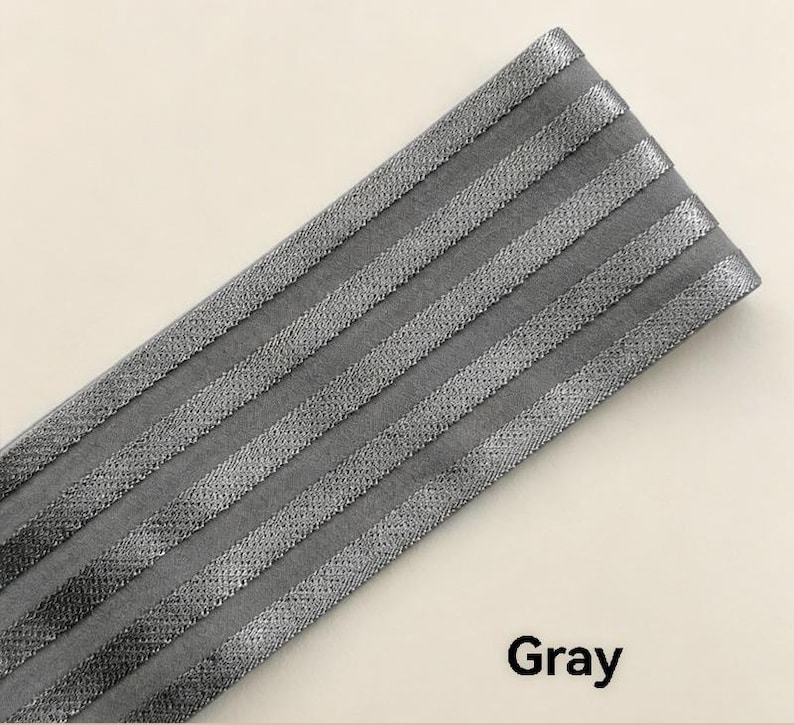 Peut inclure: Une large bande de tissu &eacute;lastique gris pr&eacute;sentant cinq rayures parall&egrave;les en relief &agrave; texture m&eacute;tallique dispos&eacute;es en diagonale. Le mat&eacute;riau poss&egrave;de une base grise mate avec une finition tiss&eacute;e scintillante sur les sections en relief et le mot Gray.